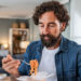 Bearded man in a denim shirt eating orange noodles from a white takeout box with a fork in a cozy kitchen setting.